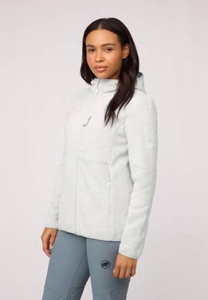 Hellgraue Fleecejacke mit hohem Kragen, vorderen Reißverschlusstaschen und einem Emblem am Ärmel. Getragen mit grauen Sporthosen.