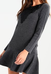Abito lungo a maniche lunghe grigio scuro con corpino aderente e gonna svasata, caratterizzato da un dettaglio in pizzo sullo scollo e un accento decorativo a forma di fiocco.