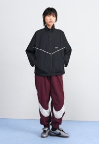 Svart vindjakke med hvite detaljer, paret med maroon joggebukser med hvite striper. Modellen har på seg grå treningssko.