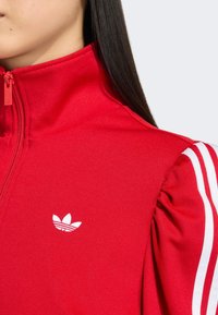 Červená športová bunda s vysokým golierom, so zipsom vpredu a bielymi trojprúžkami na rukávoch. Logo Adidas v bielej farbe na hrudi.
