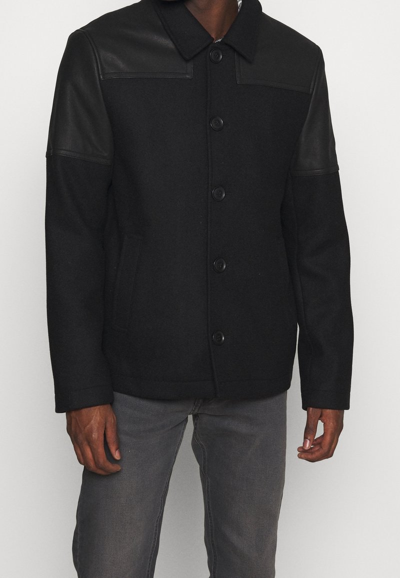 Svart ullblandad rock med läderaxlar, stora framknappar och sidofickor. Har en krage och en strukturerad passform.