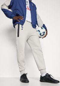 Lysegrå joggers med tætsiddende pasform, sort sidelås og ribbestrikkede manchetter, parret med en marineblå jakke og sorte sneakers, mens der holdes en fodbold.