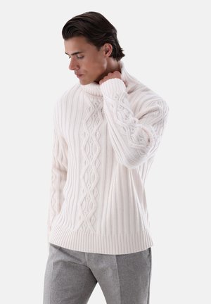 Mann trägt einen weißen kabelgestrickten Rollkragenpullover und graue Hose, schaut nach unten und berührt mit einer Hand den Kragen des Pullovers.
