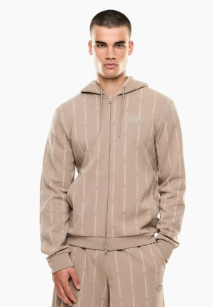 Junger Mann trägt einen beigen EA7 gemusterten Kapuzenpullover mit Reißverschluss und passende Jogginghose, steht mit einer Hand in der Tasche vor weißem Hintergrund.
