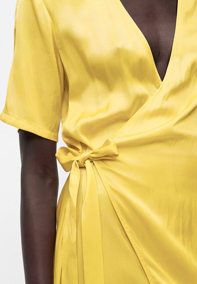 Άτομο με σκούρο δέρμα που φοράει κίτρινο σατέν φόρεμα τύπου wrap δεμένο με φιόγκο στη μέση, με κοντά μανίκια και V-λαιμόκοψη.