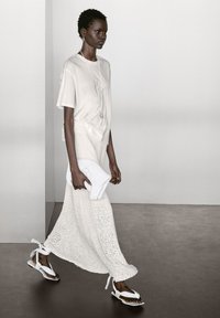 Weißes Kurzarm-Oberteil, kombiniert mit einem langen, gestrickten, cremefarbenen Rock. Sie hält eine strukturierte weiße Clutch und trägt weiße Sandalen mit Bändern.