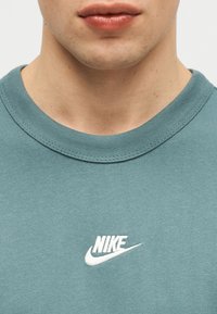 Κοντινό πλάνο του κάτω μέρους του προσώπου και του λαιμού ενός ατόμου που φοράει μια πετρόλ μπλούζα Nike με λευκό λογότυπο Nike στο κέντρο του στήθους.