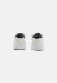 Zapatillas blancas de cuero texturizado, con un acento en el talón negro, suela resistente y un diseño minimalista. La vista trasera resalta los colores en contraste.