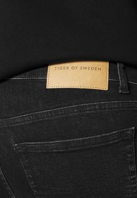 Vaqueros de mezclilla negros con una etiqueta de cuero que dice "TIGER OF SWEDEN". Las características incluyen bolsillos, costuras sutiles y una textura suave.