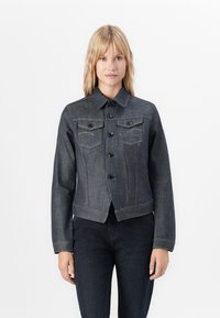 Woman with blonde hair wearing a dark denim button-up jacket and matching dark denim jeans, standing against a plain white background.