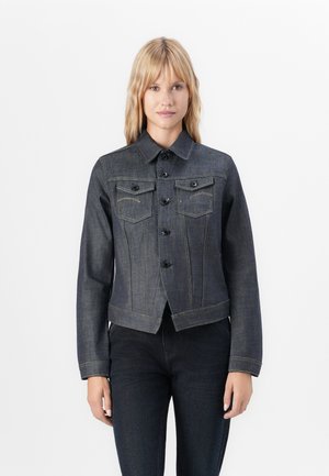 Woman with blonde hair wearing a dark denim button-up jacket and matching dark denim jeans, standing against a plain white background.