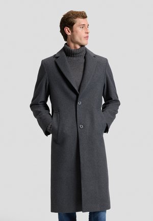Jeune homme portant un long manteau gris foncé et un pull à col roulé gris, debout les mains dans les poches, regardant sur le côté.