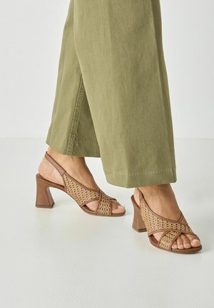 Brune sandaler med blokhæl og vævet tekstiloverdel, åbent tå-design og krydsede stropper, kombineret med olivengrønne bukser med vide ben.