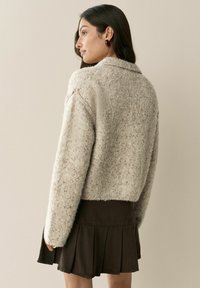 Žena s dlhými tmavými vlasmi, oblečená v bordových náušniciach, béžovom textúrovanom pletenom svetri a tmavej plisovanej sukni, stojí bokom pred jednoduchou stenou.