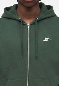 Άτομο που φοράει πράσινο φούτερ Nike με φερμουάρ, κορδόνια και λευκό λογότυπο Nike στο αριστερό στήθος.