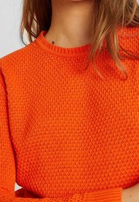 Pull orange en tricot avec un motif texturé, encolure arrondie et poignets côtelés. Le tissu semble doux et léger.