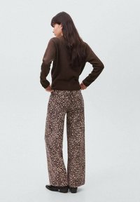 Femeie cu păr lung și închis la culoare, îmbrăcată într-un pulover maro și pantaloni larghi cu imprimeu leopard, stând cu mâinile pe șolduri, întoarsă cu spatele.