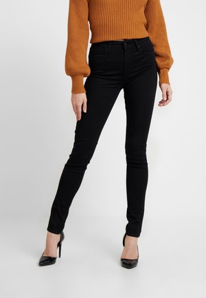 Una modelo lleva un suéter naranja acanalado y jeans ajustados negros, de pie con tacones altos negros frente a un fondo blanco.