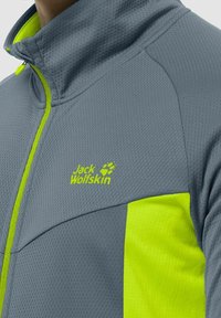Veste grise à fermeture éclair avec une surface texturée. Présente un accent jaune vif sur un côté et le logo "Jack Wolfskin" en jaune.