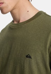Olive green cotton sweatshirt featuring a round neckline, speckled texture with yellow accents, and a small black logo on the chest.