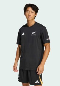 Czarna sportowa koszulka z okrągłym dekoltem, z białymi logo i wzorem pióra. Połączona z czarnymi szortami i żółtymi akcentami.