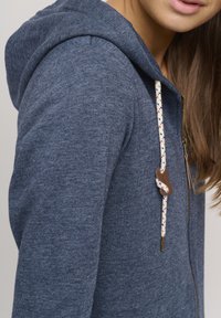 Blå fleece hettegenser med snor og flerfargede aksenter samt lærstopper. Glidelås lukking og teksturert stoff.