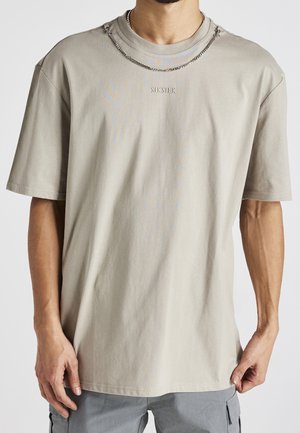 T-shirt beige clair à manches courtes en coton avec un col rond et un logo "SikSilk" brodé. Porté avec un collier en chaîne argenté.