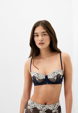 Vrouw met lang bruin haar draagt een marineblauwe en witte kanten bh en bijpassend ondergoed, staand tegen een effen witte achtergrond.