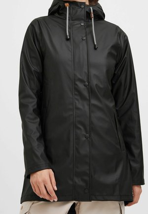 Imperméable noir à capuche avec boutons-pression à l'avant, cordons blancs et noirs, et poches latérales porté sur un pantalon clair.