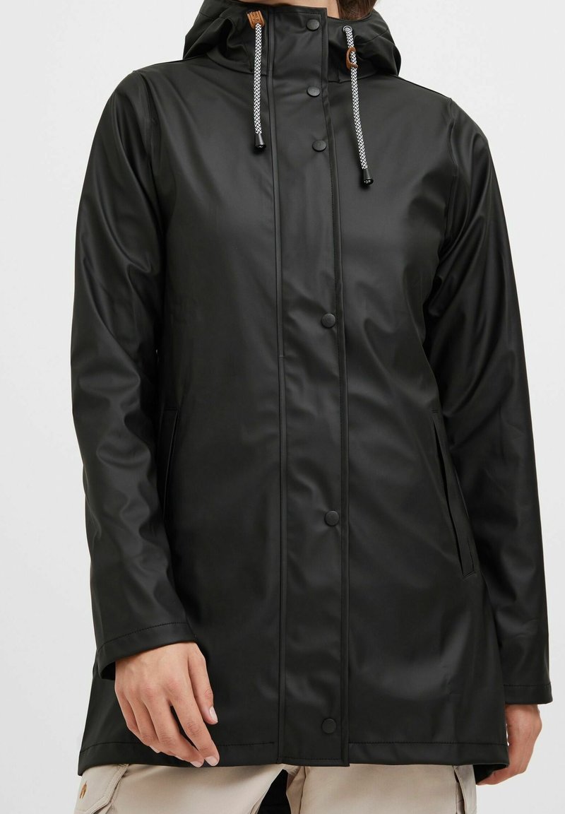 Imperméable noir à capuche avec boutons-pression à l'avant, cordons blancs et noirs, et poches latérales porté sur un pantalon clair.