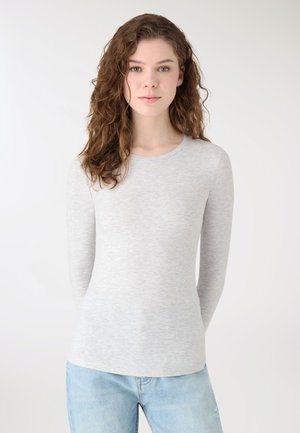 Młoda kobieta o kręconych włosach, ubrana w jasnoszarą bluzkę z długim rękawem i jasnoniebieskie dżinsy, stojąca na jednolitym białym tle.