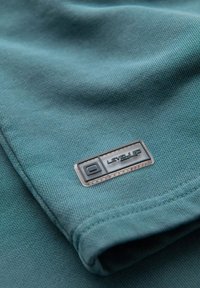 Chemise en tissu teal avec une texture lisse, comportant une étiquette rectangulaire noire portant l'inscription "LEVEL UP" en texte blanc.