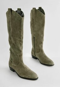 Botas altas de ante verde oliva con puntera puntiaguda, costuras decorativas, cremallera lateral y tacón bajo tipo bloque. Textura suave en toda la bota.