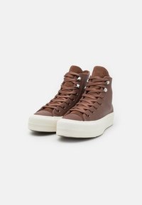 Brązowe sneakersy wysokie, wykonane z teksturowanego materiału, z białą gumową podeszwą, metalowymi oczkami i pasującymi brązowymi sznurówkami.
