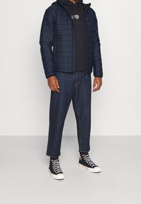 Marineblauwe gewatteerde jas met capuchon, gedragen over een zwarte hoodie, gecombineerd met losse donkere jeans en zwarte hoge sneakers.