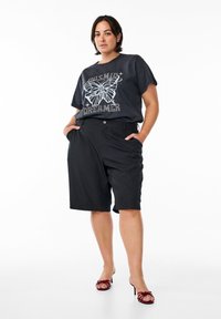 Černé grafické tričko má bílé nové design motýla s nápisem "DREAMER". Kombinováno s černými šortkami ke kolenům a burgundskými sandály na otevřené špičce.