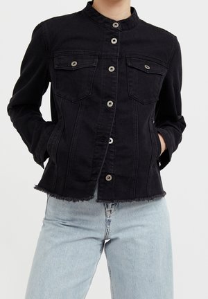 Person trägt eine schwarze Jeansjacke mit Knöpfen und Brusttaschen sowie hellblaue Jeans, die Hände in den Jackentaschen, steht vor weißem Hintergrund.