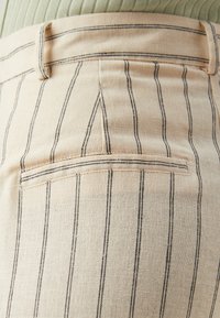 Beige fabric pants with vertical black stripes, featuring a back pocket and a waistband, highlighting textured material and tailored design.