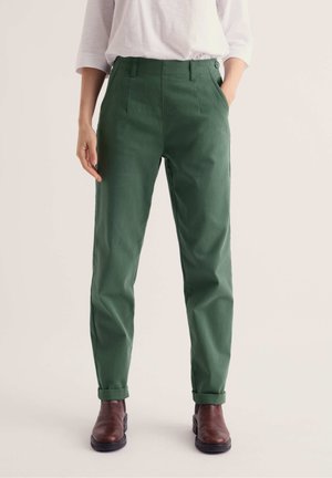Femme portant un pantalon vert taille haute fuselé avec ourlets retroussés, un haut blanc à manches longues et des bottines marron, debout les mains dans les poches.