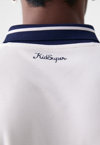 Bijela polo majica s teksturiranim materijalom, s tamnoplavim ovratnikom s dva bijela pruga i izvezenim logom "KidSuper" u tamnoplavo.