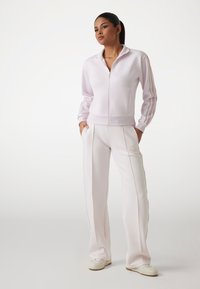 Veste zippée rose clair avec col montant et poches avant, associée à un pantalon assorti présentant des rayures sur les côtés et des détails de logo brodés.