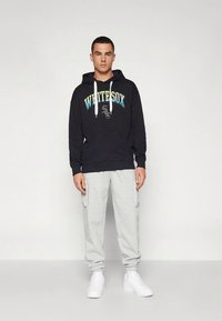 Svart hettegenser med "WHITE SOX"-grafikk i lyseblått og gult, kombinert med lysegrå joggebukser og hvite joggesko.