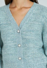 Cardigan tricotat de culoare bleu deschis, cu decolteu în V, patru nasturi din perlă și un material texturat cu accente din fir de aur.