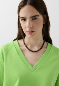 Pulover cu decolteu în V, de culoare verde aprins, din bumbac, cu textură ribbed și o croială lejeră, asortat cu un colier din mărgele negre, netede.