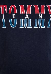Námořnická modrá tričko s výrazným nápisem "TOMMY JEANS" v červené a světle modré barvě. Tkanina se zdá být jemná s texturovaným povrchem.