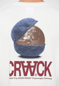 Λευκό t-shirt με γραφικό σχέδιο ενός μπλε ημισφαιρίου που αποκαλύπτει μια καφέ υφή σφαίρα, με έντονο κόκκινο κείμενο από κάτω που γράφει "CRACK."