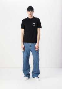 Black cotton t-shirt with white logo on the chest, paired with blue cargo jeans featuring large side pockets and white sneakers. Black cap worn.