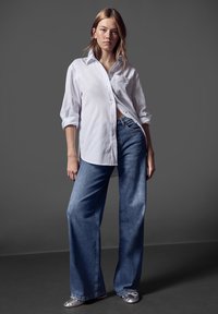 Camisa blanca de botones con las mangas arremangadas, combinada con pantalones vaqueros de pierna ancha en azul. Unos zapatos planos plateados añaden un toque metálico al conjunto.