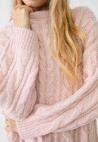 Ljust rosa kabelstickad tröja med hög hals och stora ärmar, med en mjuk textur och intrikat mönster.