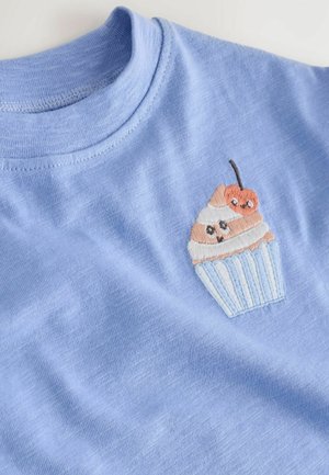 Lyseblå bomuld T-shirt med et broderet cupcake-design med creme, brune og kirsebær nuancer, sat mod en glat stoftekstur.